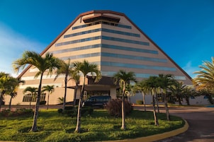 Um edifício moderno com telhado triangular e vários andares, rodeado por palmeiras e um gramado bem cuidado.