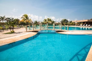 Piscina azul límpida com uma ponte de madeira, rodeada por espreguiçadeiras e palmeiras.