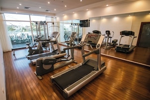 Uma academia com diversos aparelhos de cardio, uma televisão e grandes janelas.