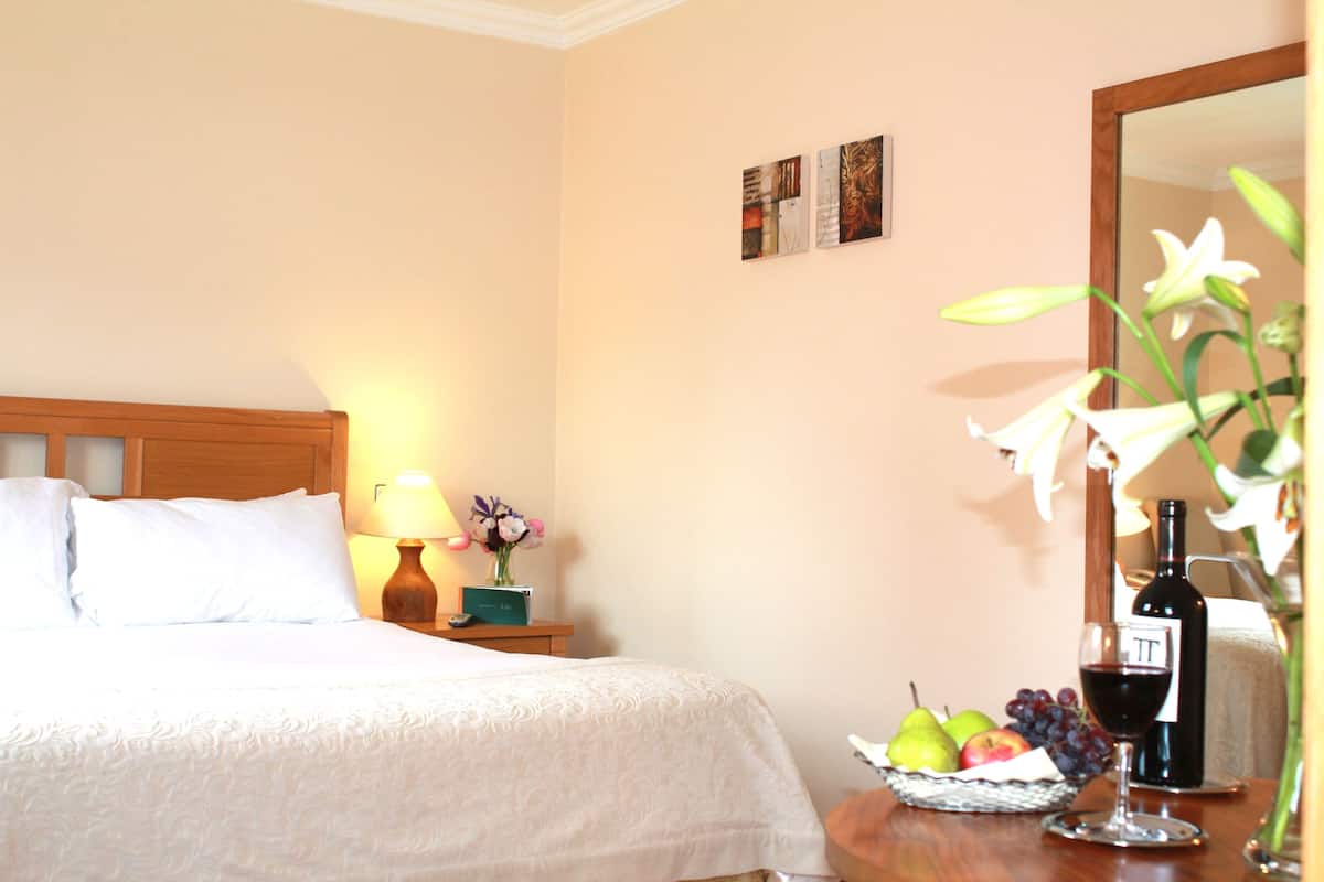 Habitación de hotel con una cama bien hecha, una mesita de noche con lámpara, un jarrón con flores, una cesta de frutas, una botella de vino y una copa de vino.