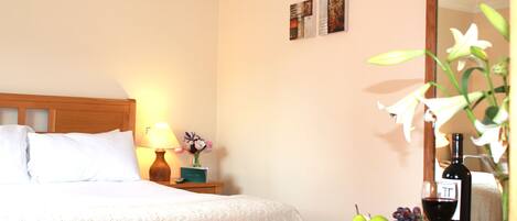 A hotel room with a neatly made bed, a nightstand with a lamp, a vase with flowers, a fruit basket, a wine bottle, and a wine glass.