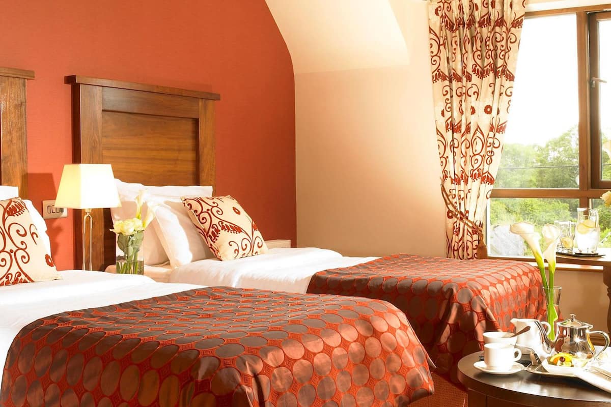 Habitación de hotel con dos camas, una ventana con cortinas, una mesita de noche con lámpara y un arreglo para el desayuno sobre la mesa.