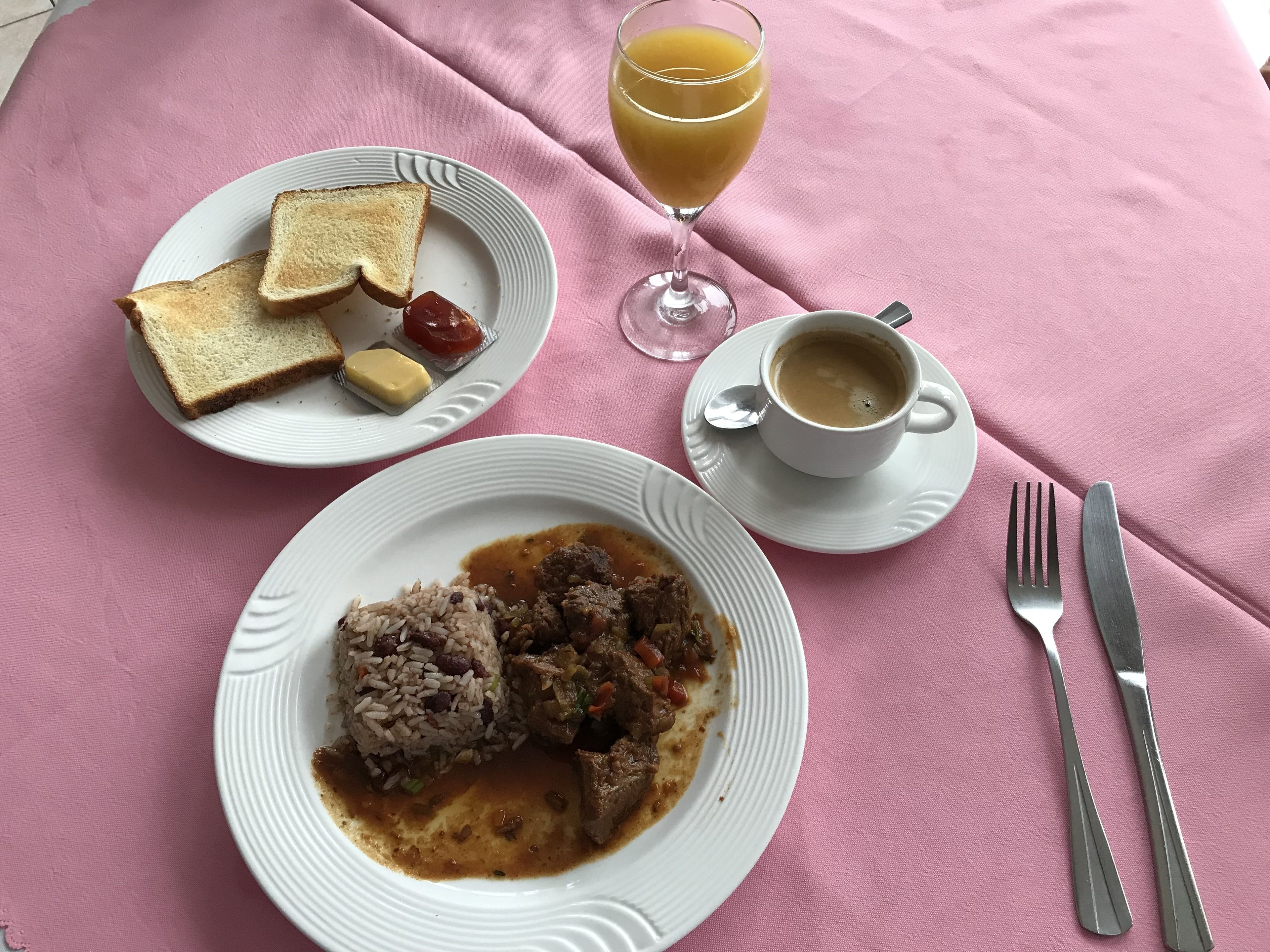 A breakfast setting with toast, butter, jam, a glass of orange juice, a cup of coffee, and a plate of meat and rice.