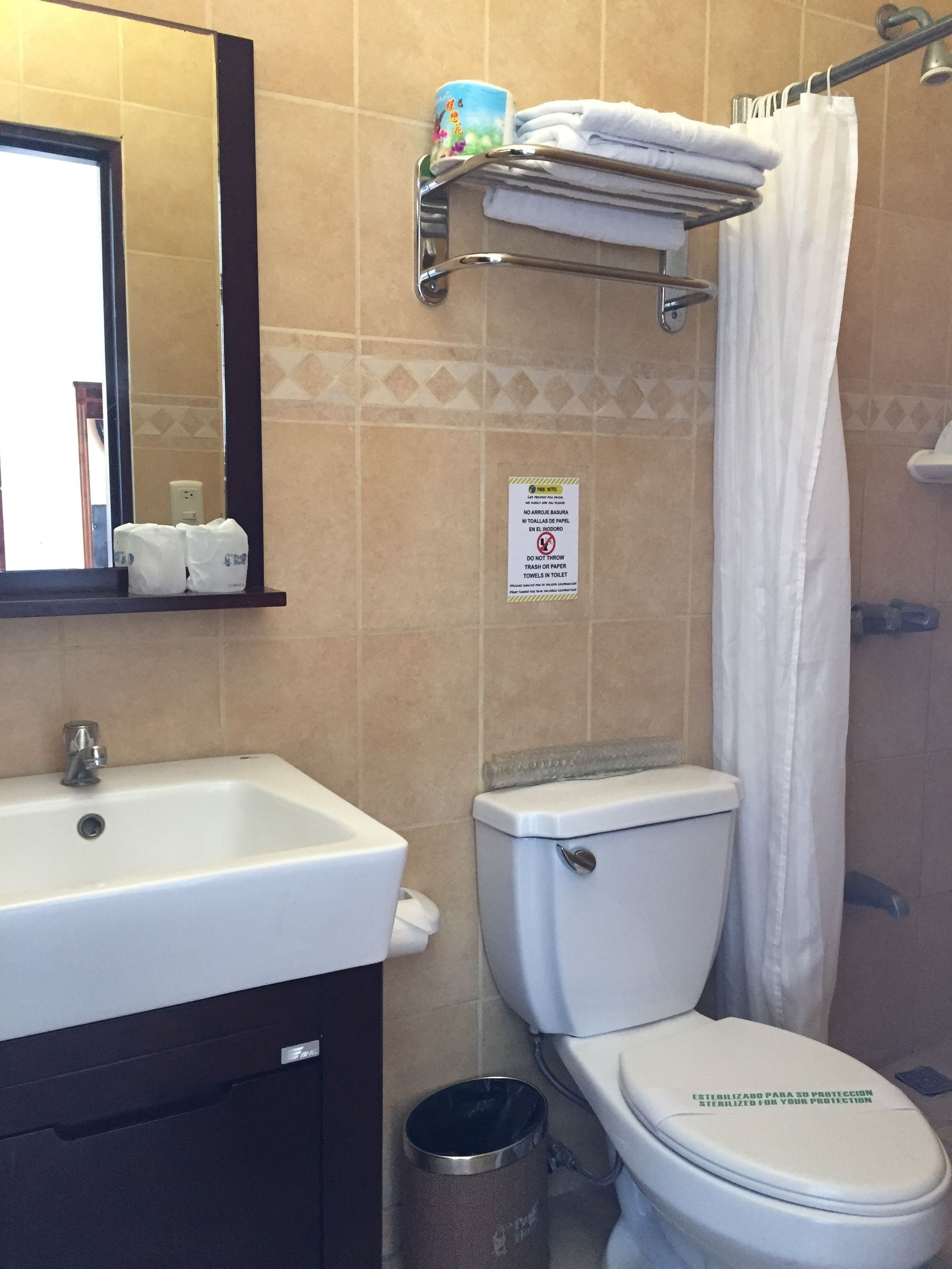 Un bagno con lavandino, WC e tenda della doccia. Si nota un cartello appeso al muro e un cestino.