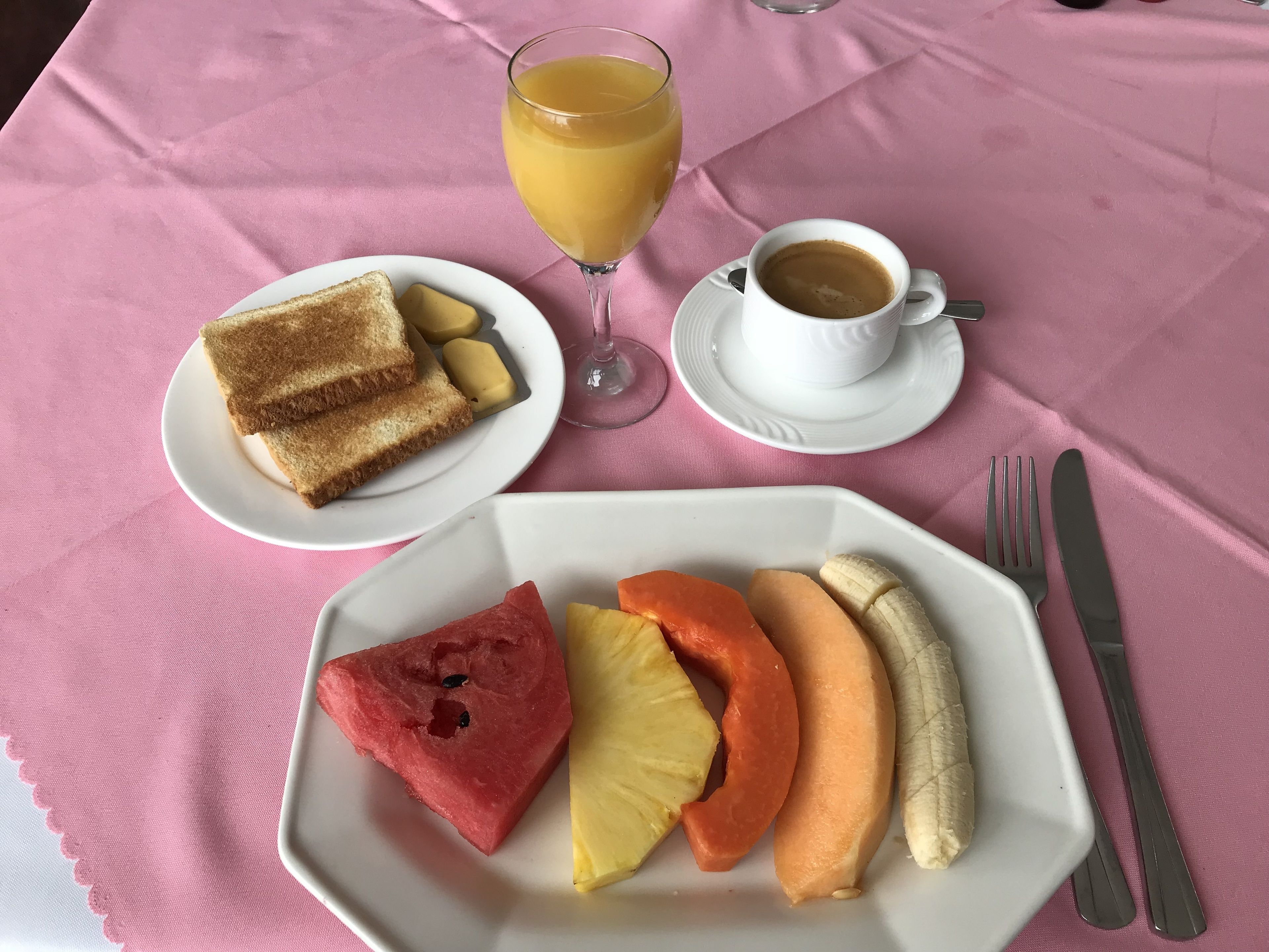 A breakfast setting with toast, butter, a glass of orange juice, a cup of coffee, and a plate of fruit including watermelon, pineapple, and bananas.