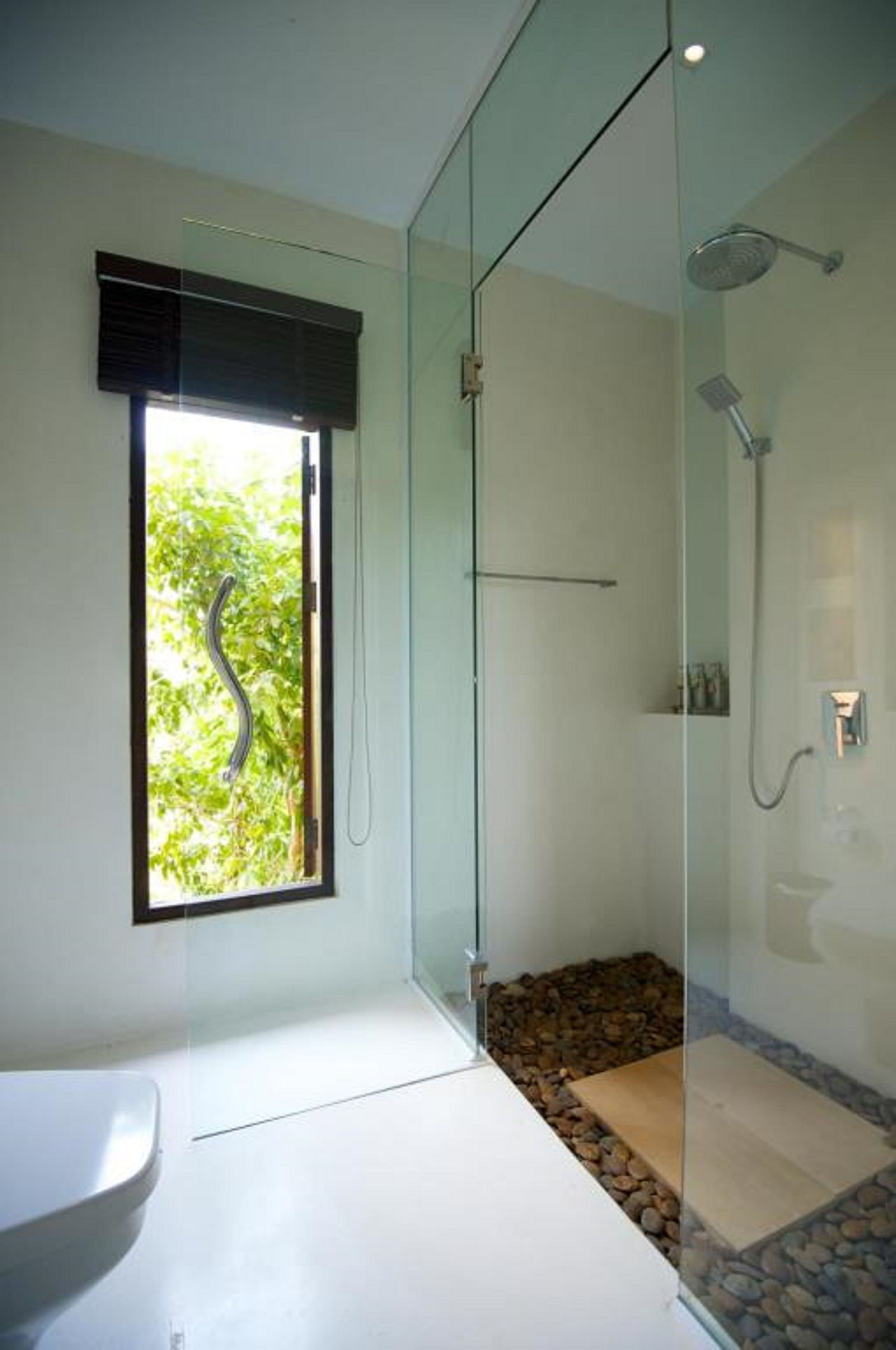 A modern bathroom with a glass shower enclosure, a white sink, and a window with a view of greenery.