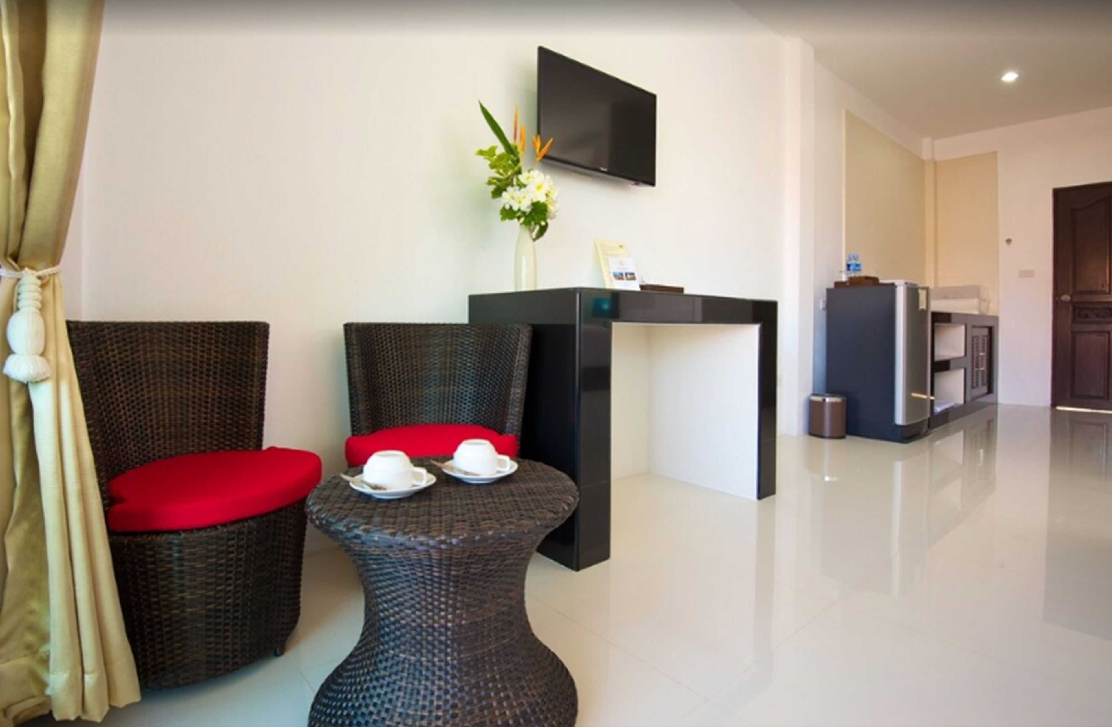 A modern hotel room with wicker furniture, a flat-screen TV, a small table with cups, and a kitchen area.