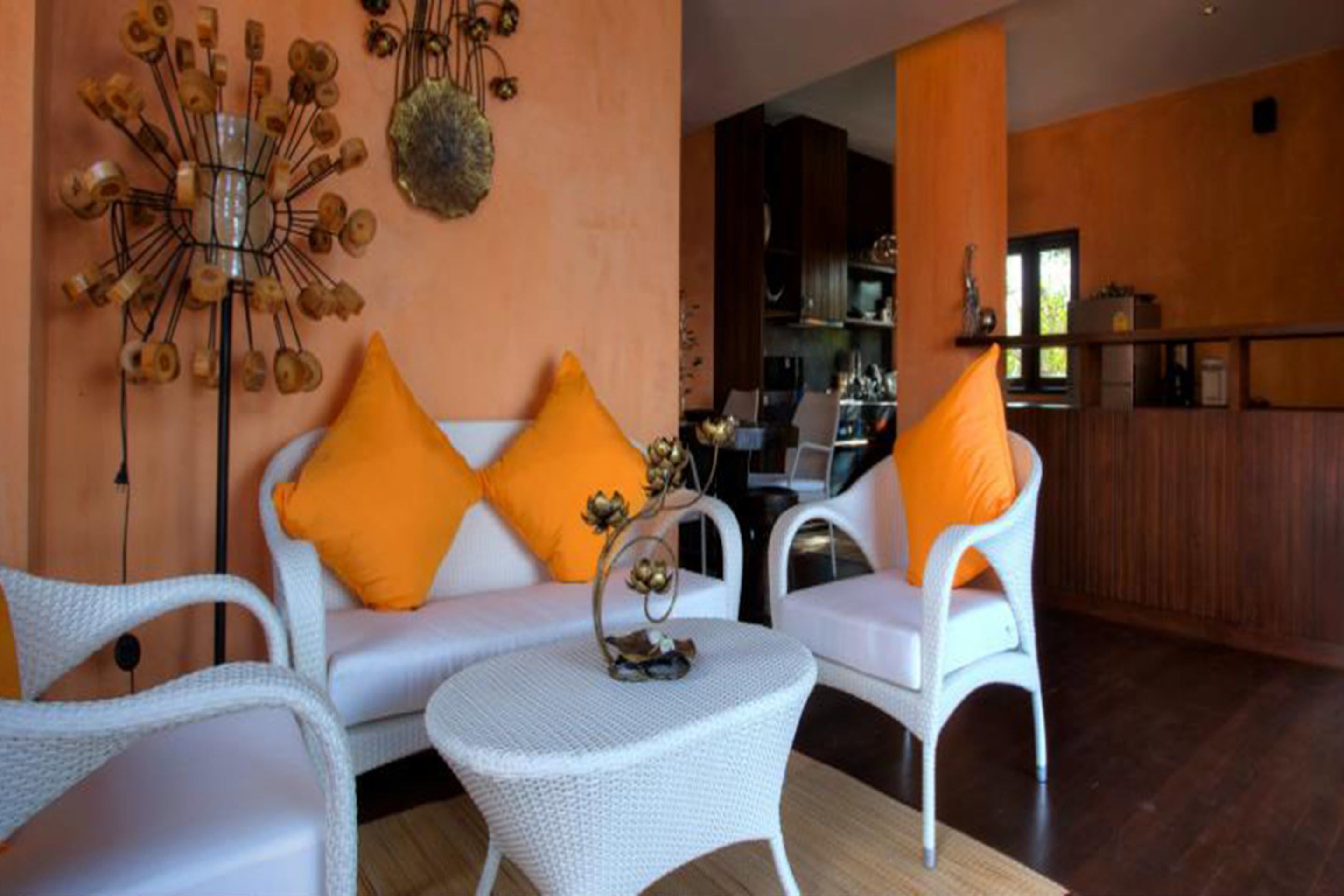 Un sofá de mimbre blanco con cojines naranja, una mesita redonda y un adorno metálico en la pared.