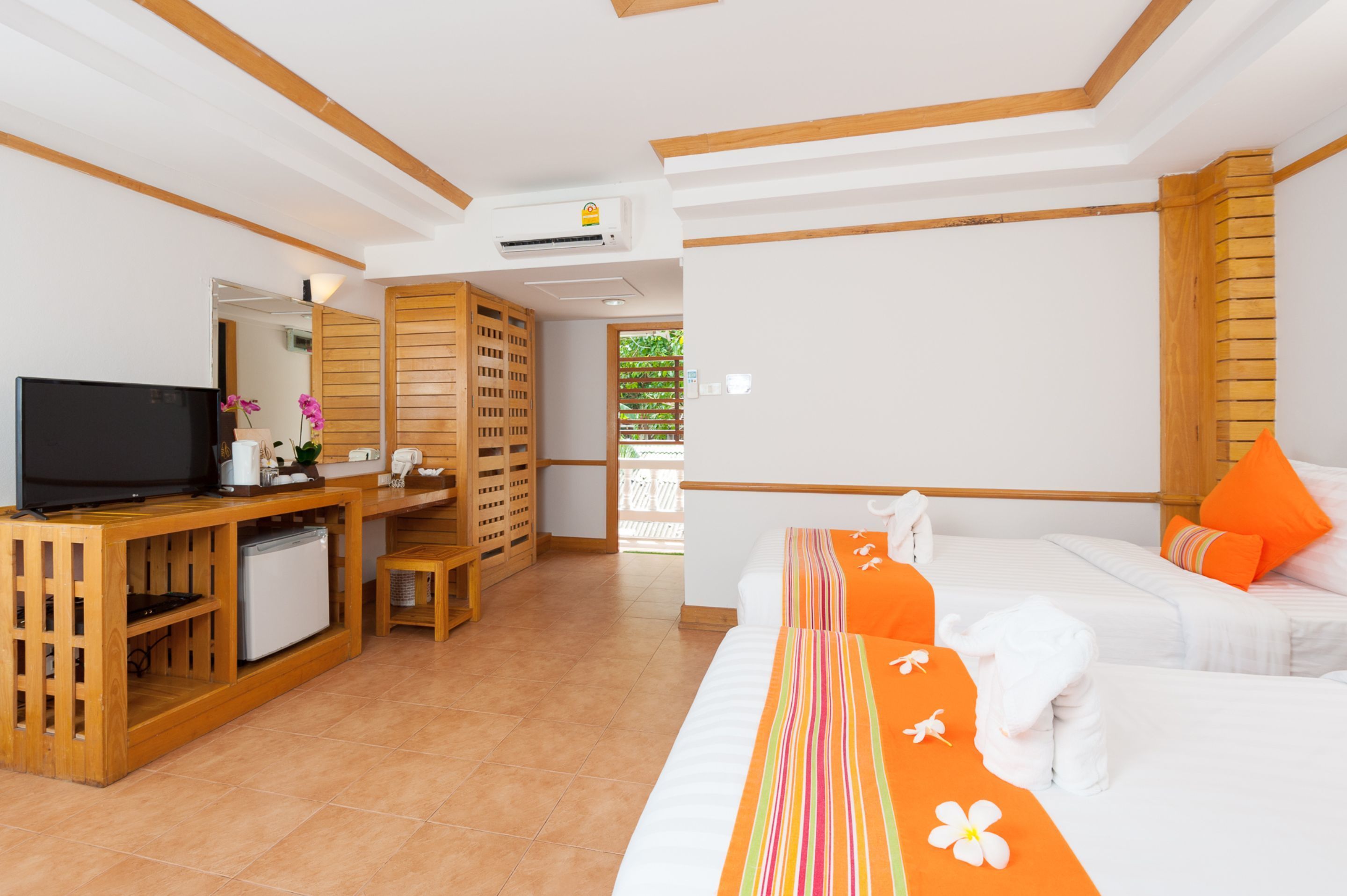 A hotel room with a TV, wooden furniture, a bed with orange pillows, and a view of greenery outside.