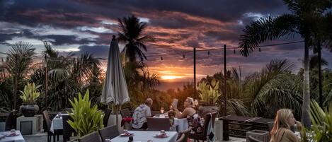 An outdoor dining area with tables set for two, surrounded by lush greenery and palm trees, under a sunset sky.