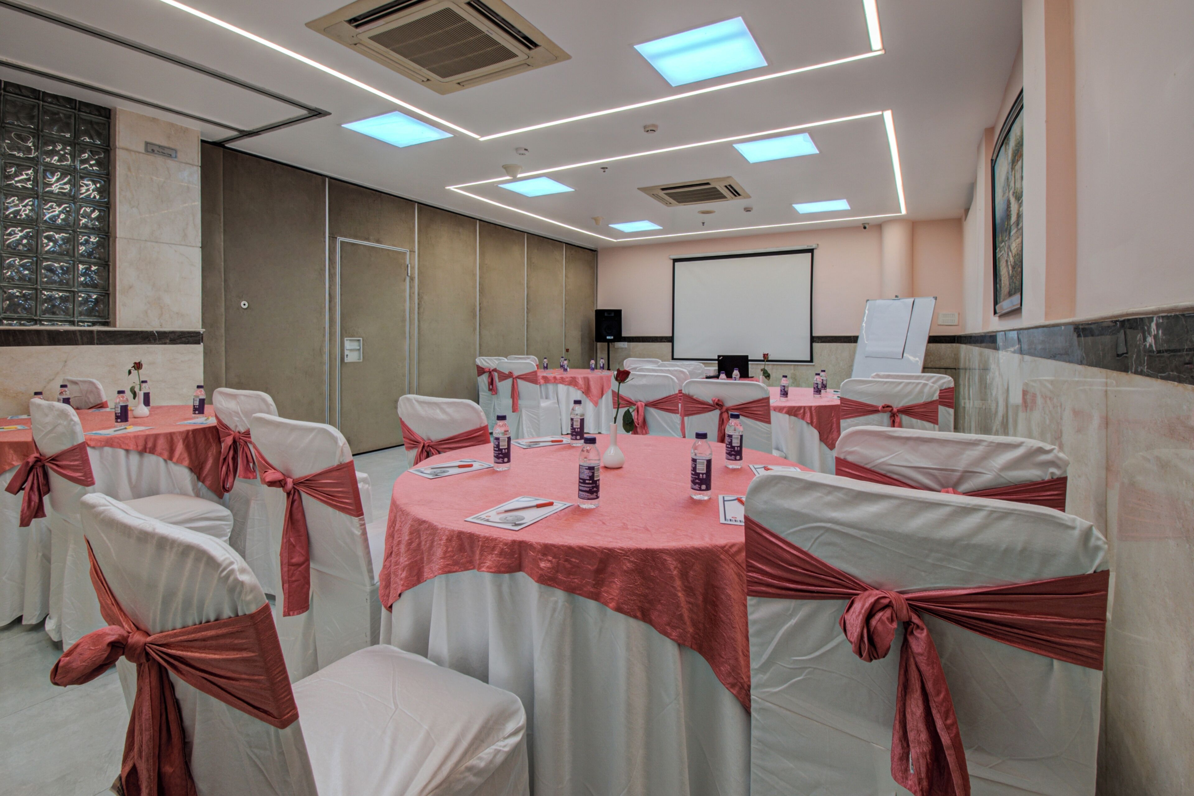 A conference room with round tables, white chairs with red sashes, a projector screen, and a whiteboard.