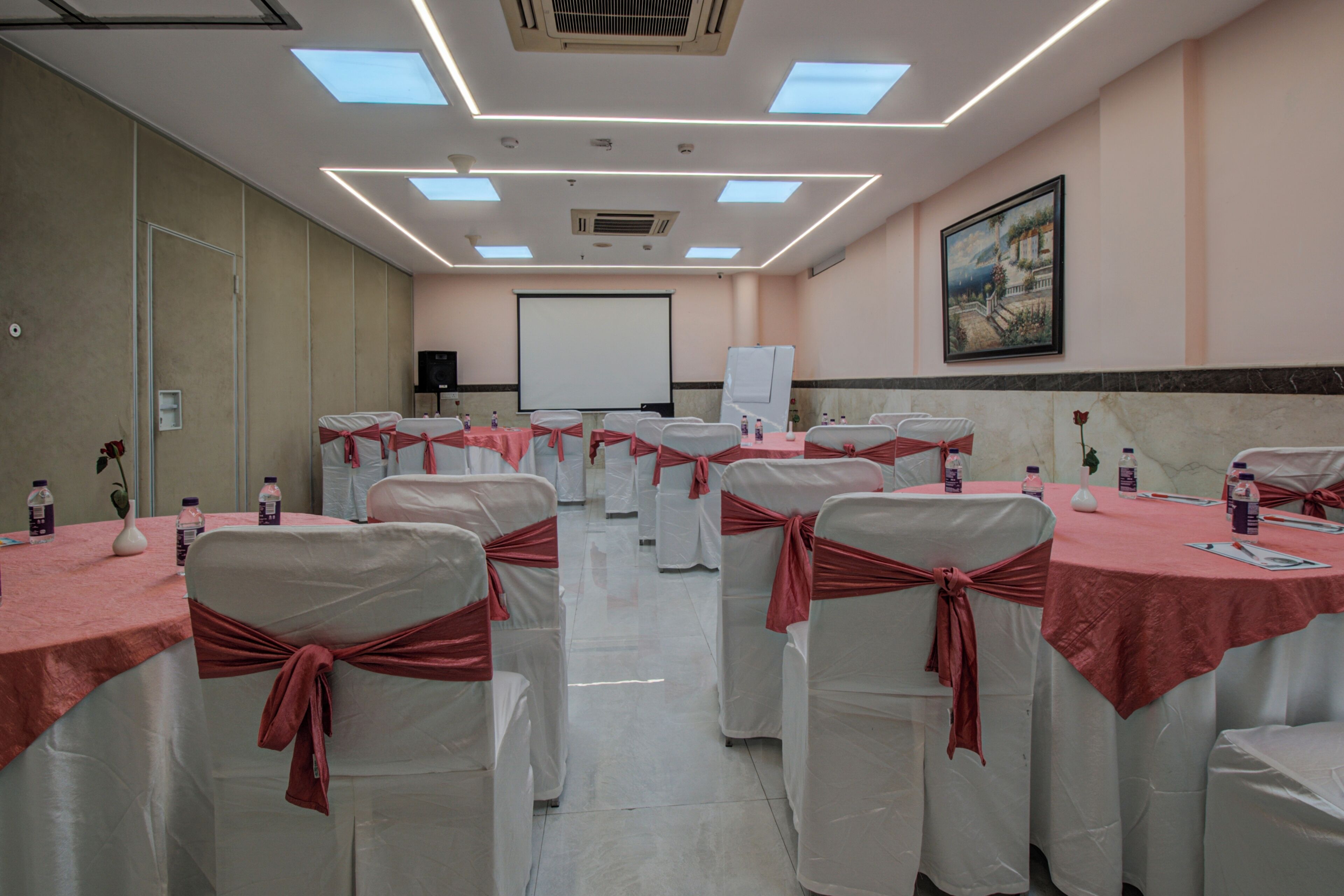 Un salón de conferencias con sillas y mesas blancas, cada una adornada con una cinta roja. Hay botellas de agua sobre las mesas, una pantalla de proyector y un cuadro en la pared.