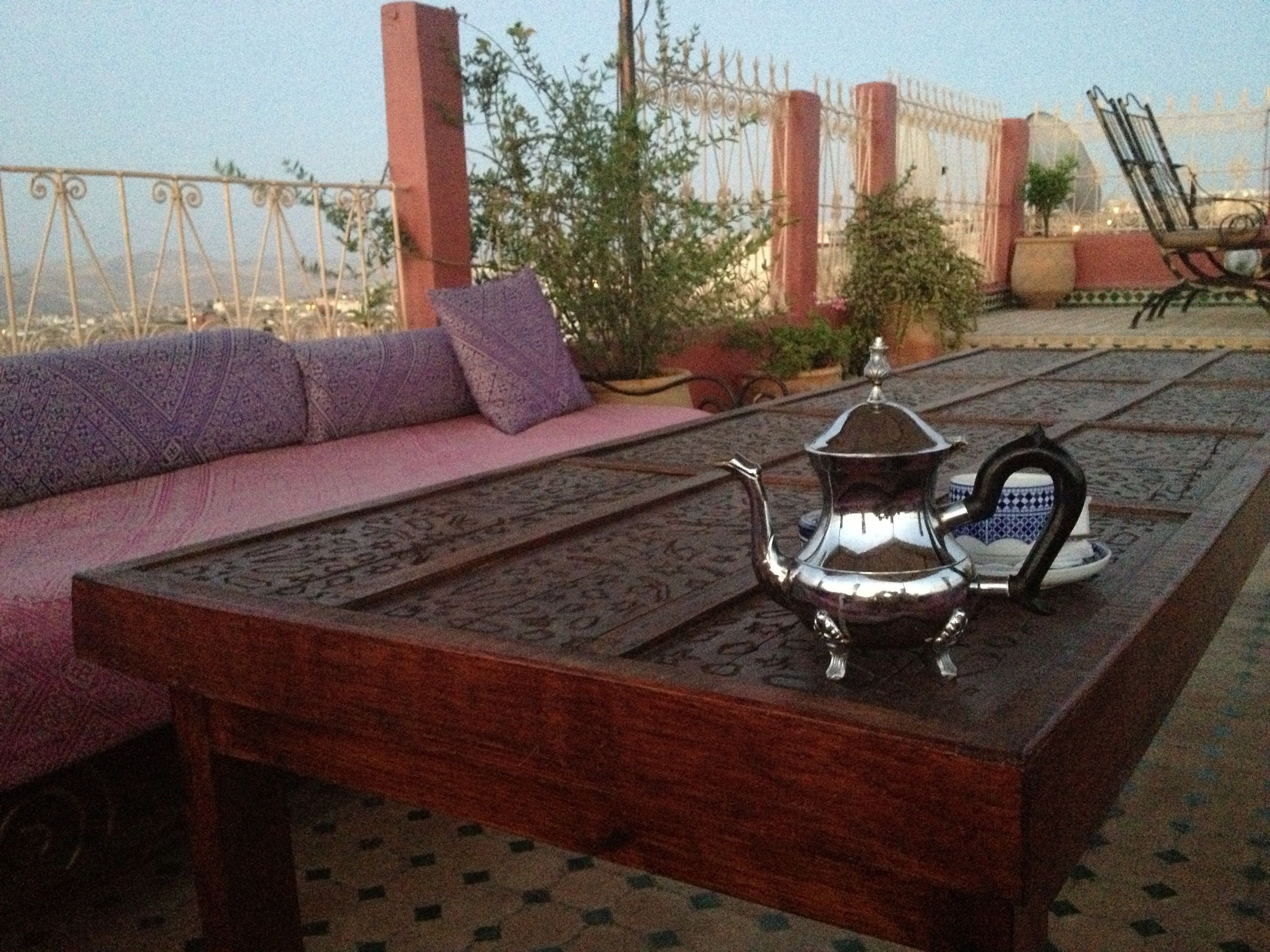A wooden table with a metal teapot and a cup on it, a pink sofa with cushions, and a metal fence in the background.