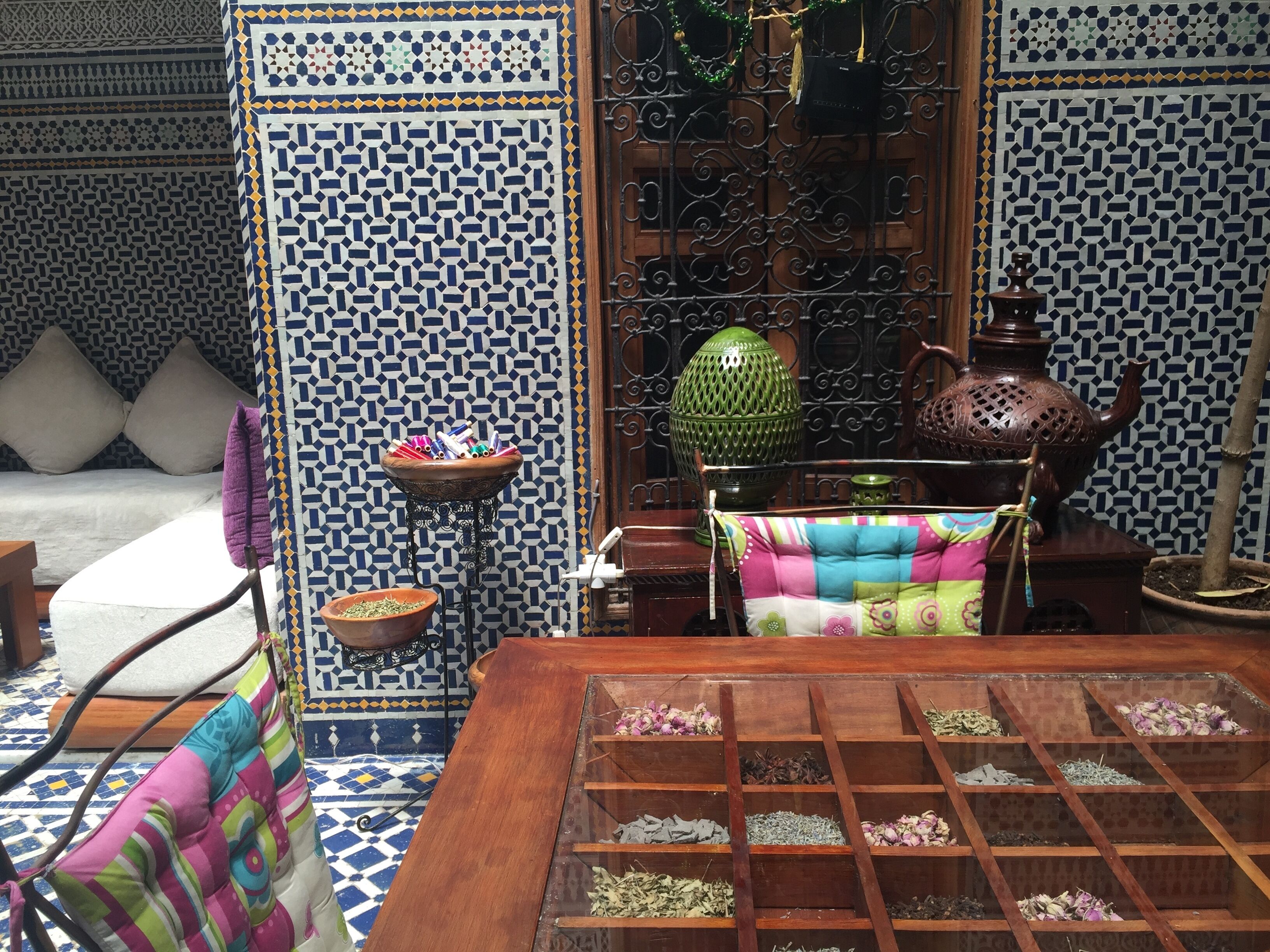A traditional Moroccan room with patterned tile walls, a wooden table with a tea set, and a colorful cushion.