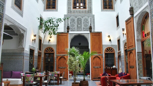 A courtyard with a central archway, surrounded by seating areas and tables, featuring traditional Moroccan tile flooring and ornate wall designs.