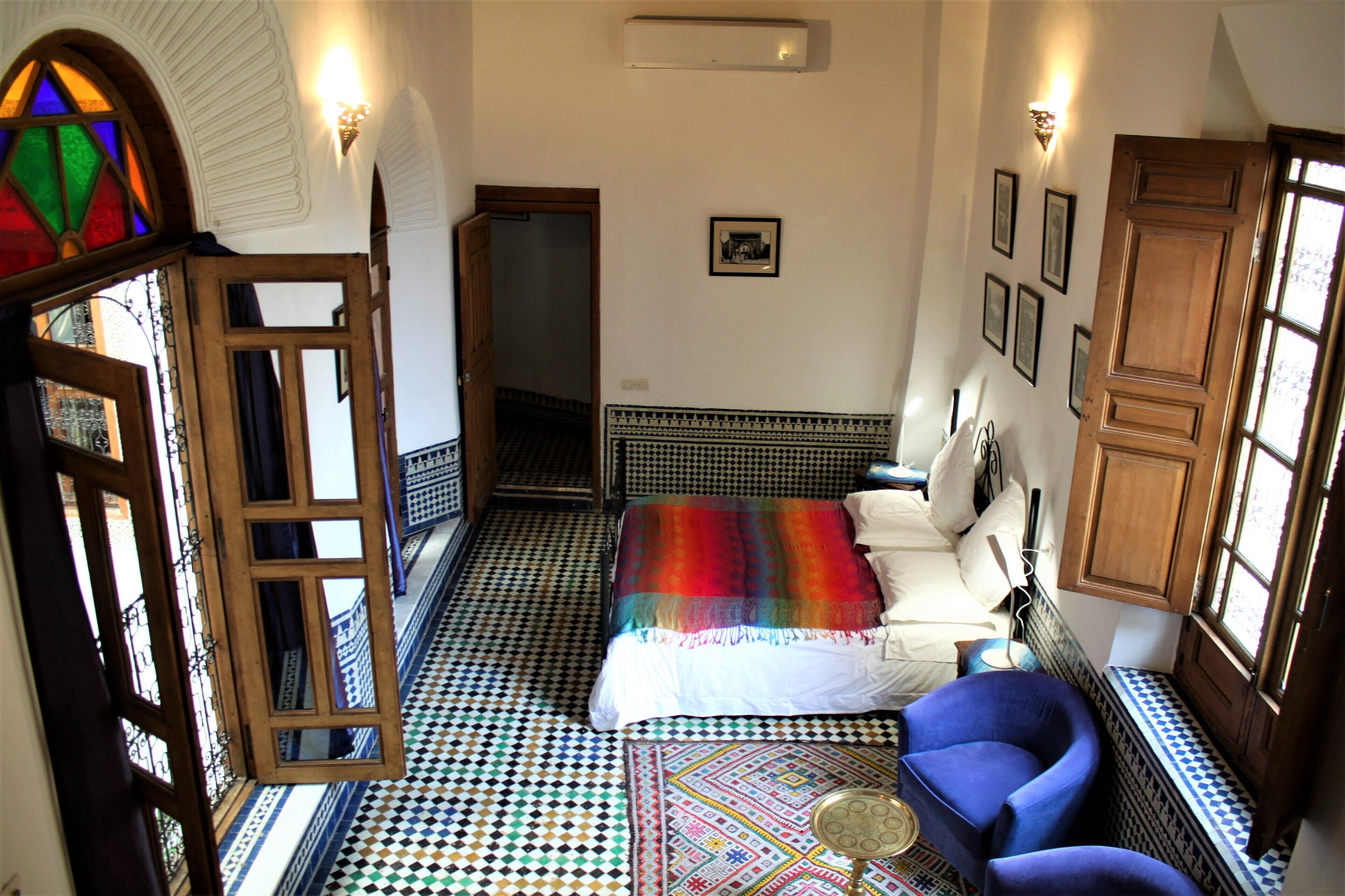 Una habitación con piso estampado, una cama con manta colorida, un sillón azul y una mesita.