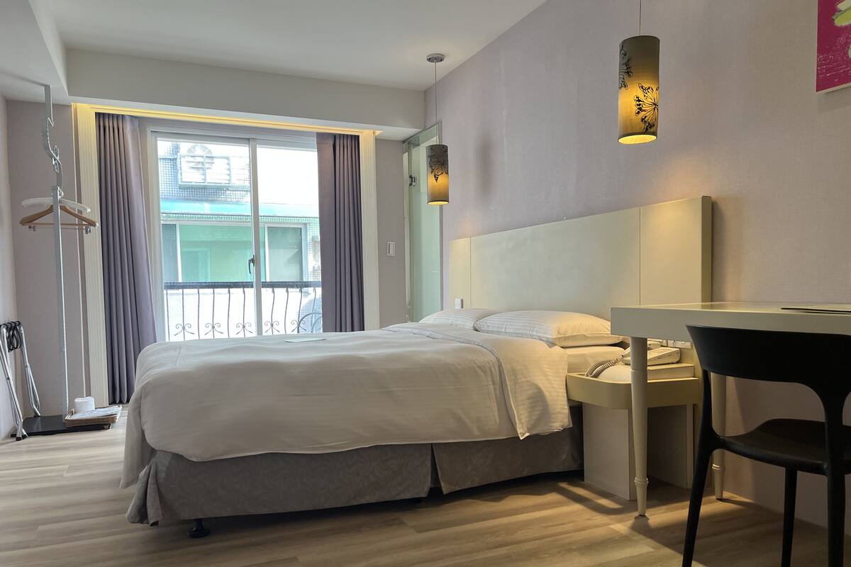 Une chambre d’hôtel moderne avec un grand lit, un bureau et une chaise. Elle dispose d’un balcon avec des rideaux et offre une vue sur l’extérieur.