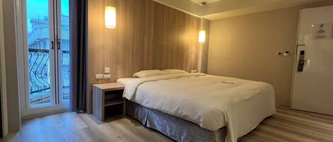 Ein modernes Hotelzimmer mit Bett, Nachttischen, einem Kleiderschrank und einem Balkon mit Blick auf Gebäude.