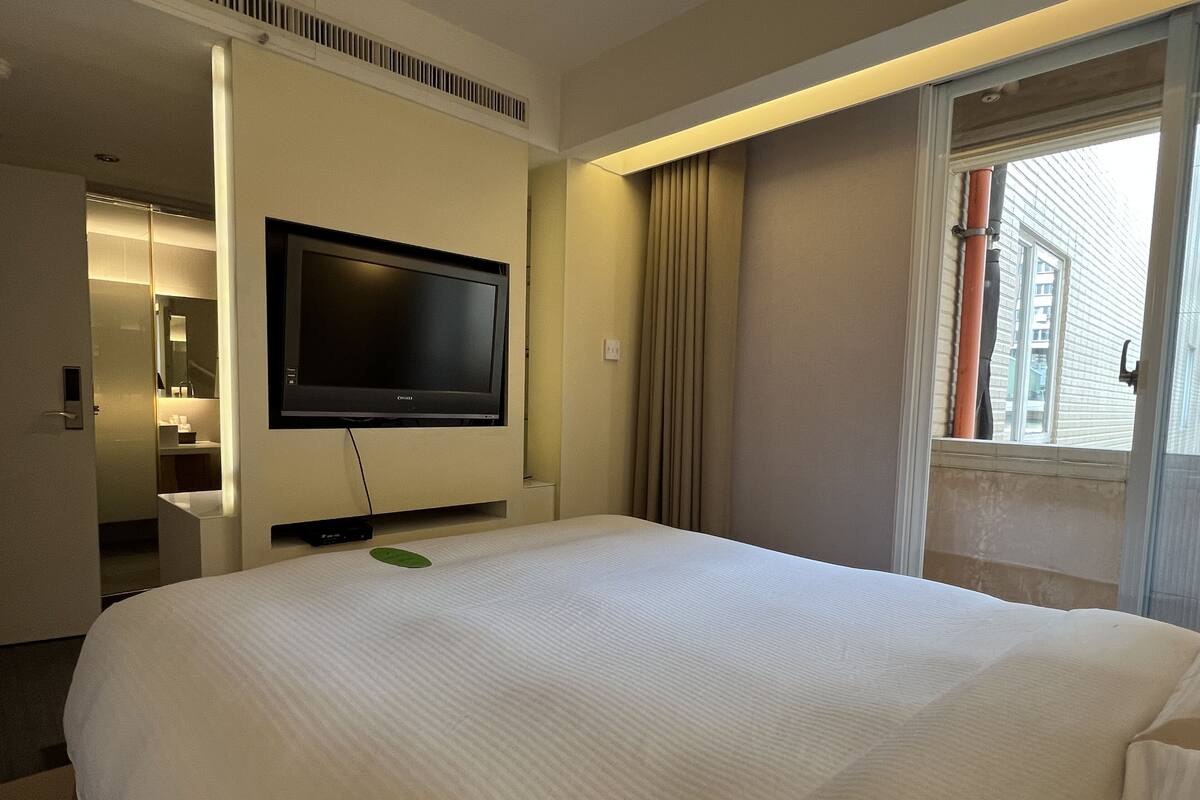 Une chambre d’hôtel avec un grand lit, un téléviseur à écran plat fixé au mur, une fenêtre avec des rideaux et une vue sur les bâtiments environnants.