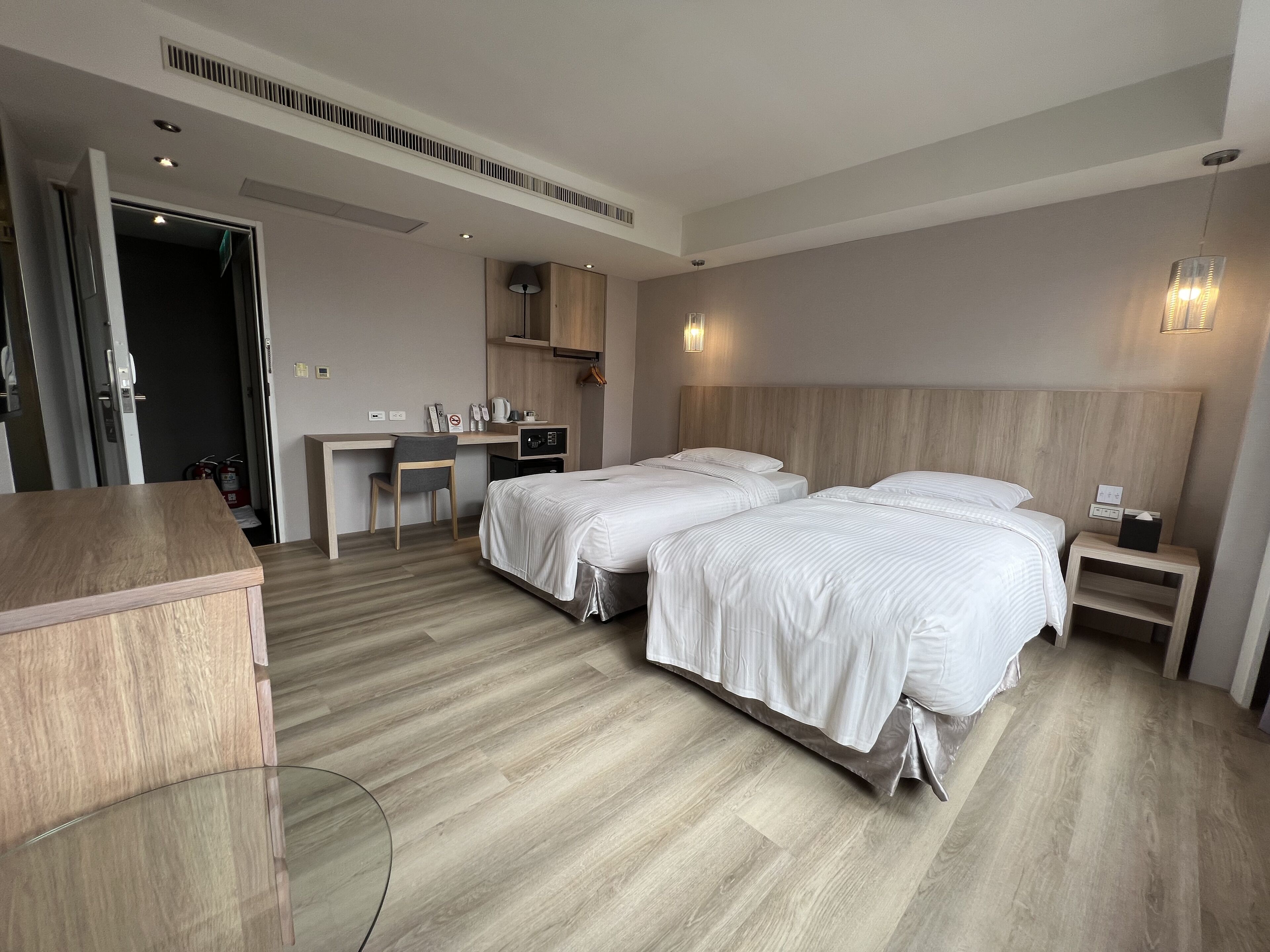 Une chambre d’hôtel avec deux lits, un bureau, une chaise, une petite table et une table de chevet avec une lampe.