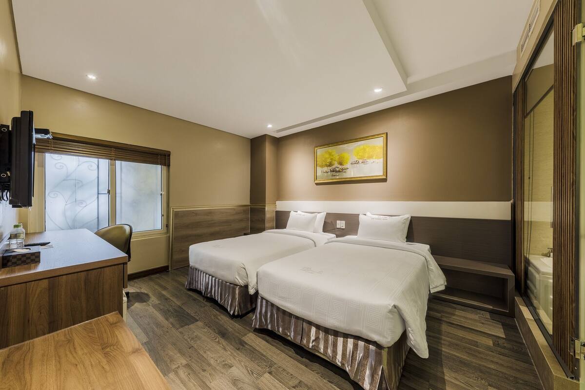 A hotel room with two beds, a desk, a window with blinds, a painting on the wall, and a wooden floor.