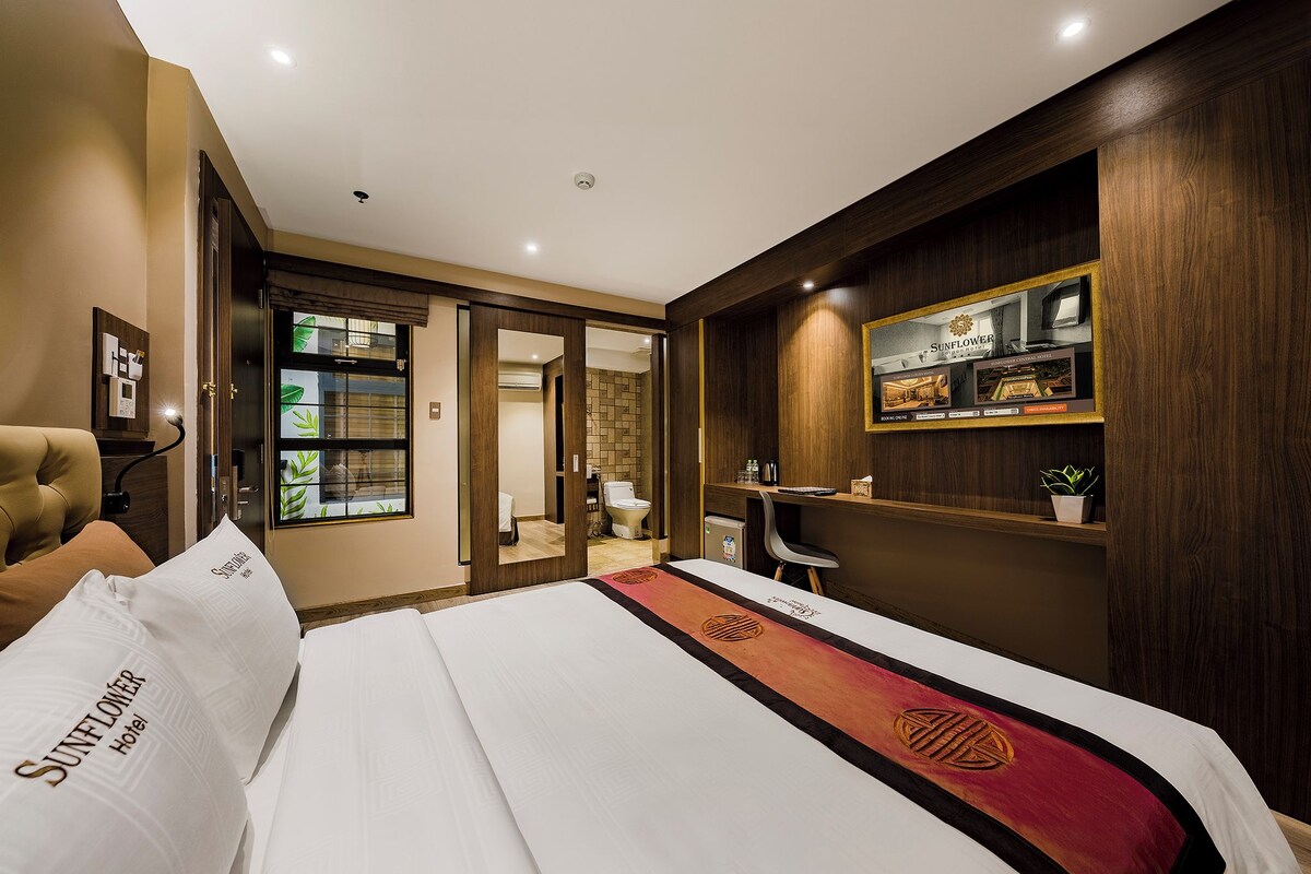Habitación de hotel con cama, escritorio, silla, estante con una planta y baño visible a través de una puerta abierta.