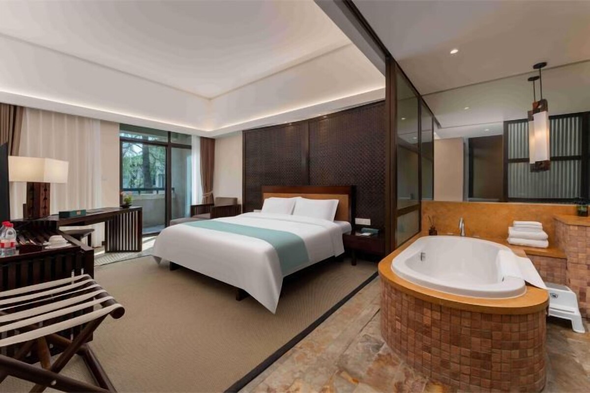 A modern hotel room with a large bed, a sitting area with a desk and chair, a bathtub, and a large mirror.