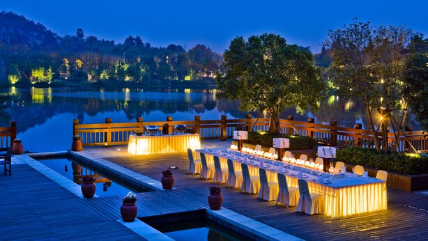 A lakeside dining area with a long table set for a meal, surrounded by chairs and potted plants. - NARADA Resort & Spa Liangzhu (Hangzhou)