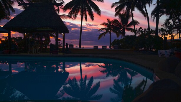 Une piscine qui reflète les palmiers et le ciel au coucher du soleil.