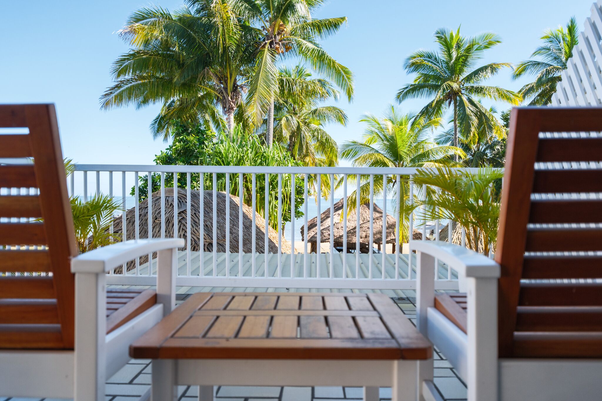 Een houten tafel en twee houten stoelen op een balkon met een witte reling, met uitzicht op palmbomen en een strand.