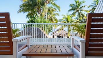 A wooden table and two wooden chairs on a balcony with a white railing, overlooking palm trees and a beach.