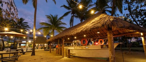 A beachside restaurant with a thatched roof, outdoor seating, and a boat moored nearby.