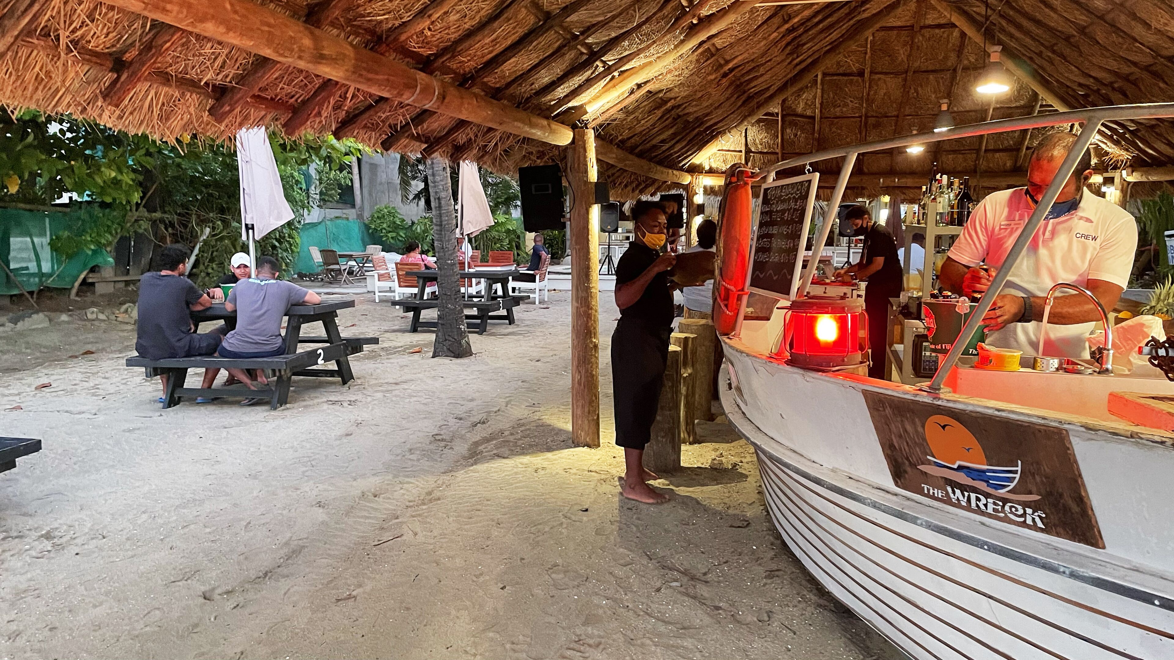 Ein Imbissstand direkt am Strand mit einem Reetdach, einer Theke mit einer Neonreklame und mehreren Tischen mit Picknickbänken, an denen Gäste essen.