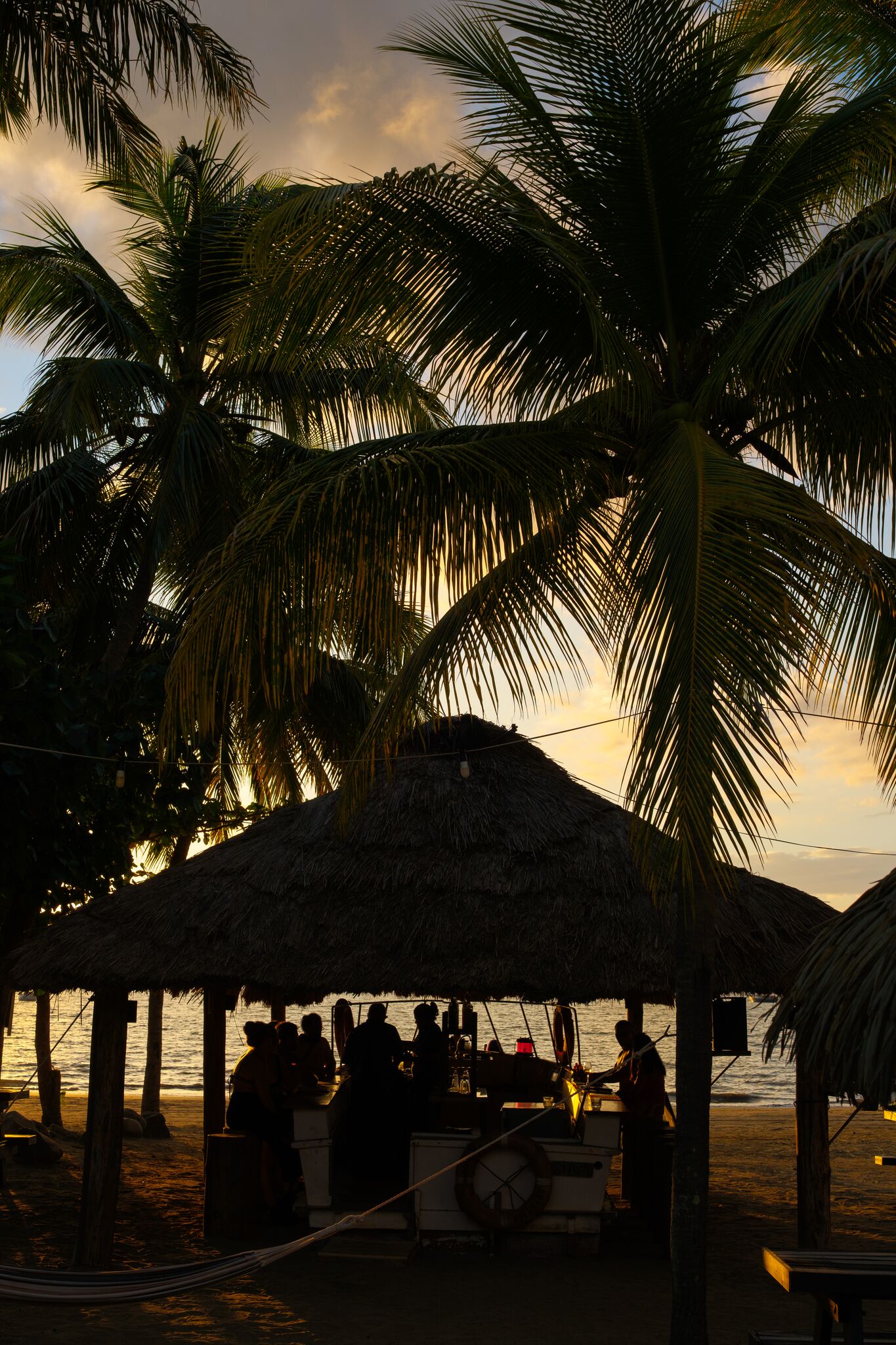 Eine Strandhütte mit einem Reetdach, Palmen und einer Gruppe von Menschen, die sich um einen Tisch versammelt haben.