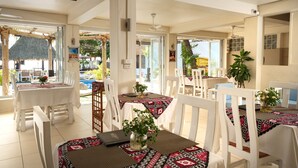 Un restaurant avec des tables et des chaises blanches, une piscine en arrière-plan et une structure surmontée d’un toit de chaume.