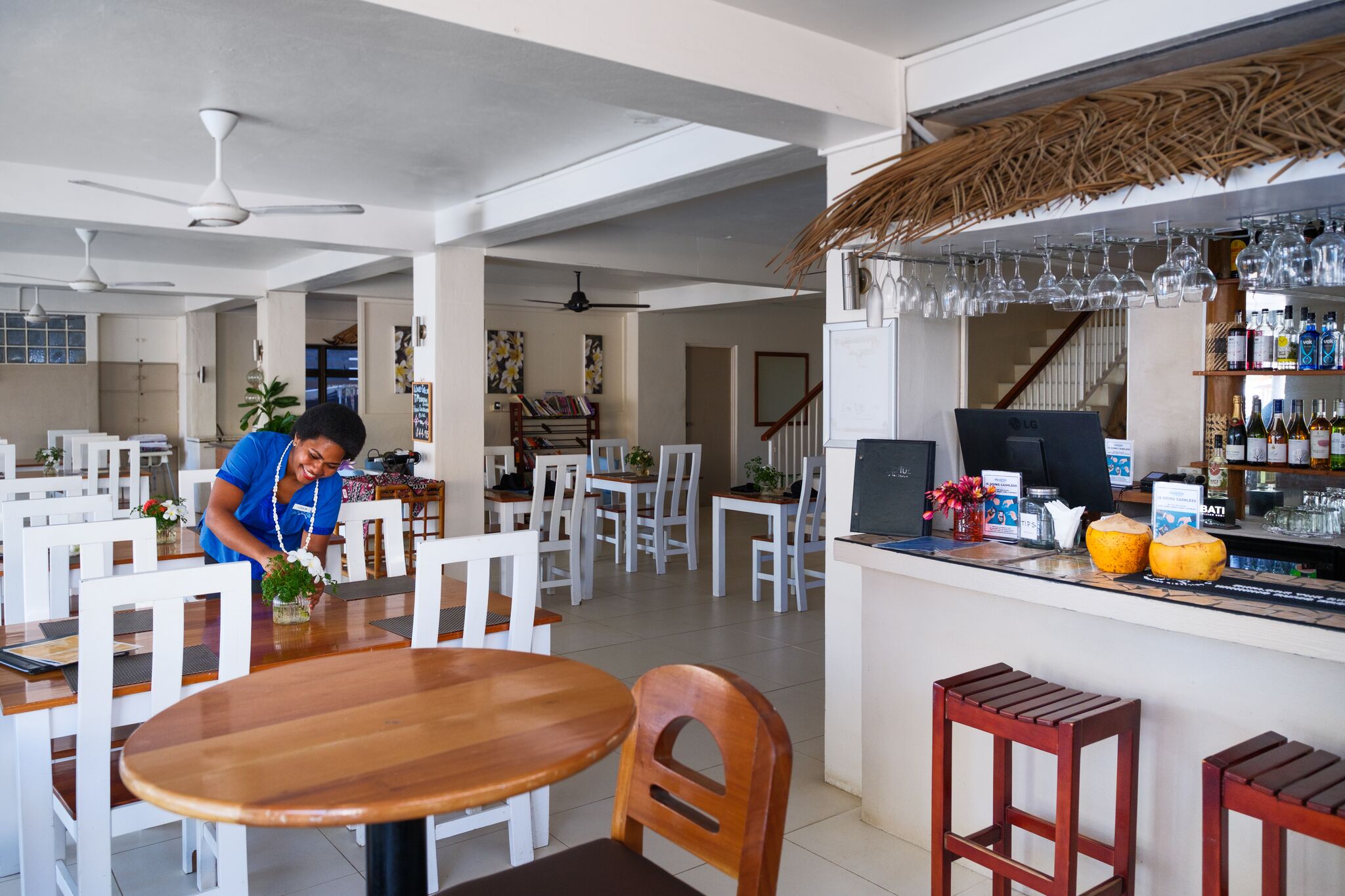 A spacious restaurant with white chairs and tables, a bar area with bottles, and ceiling fans.