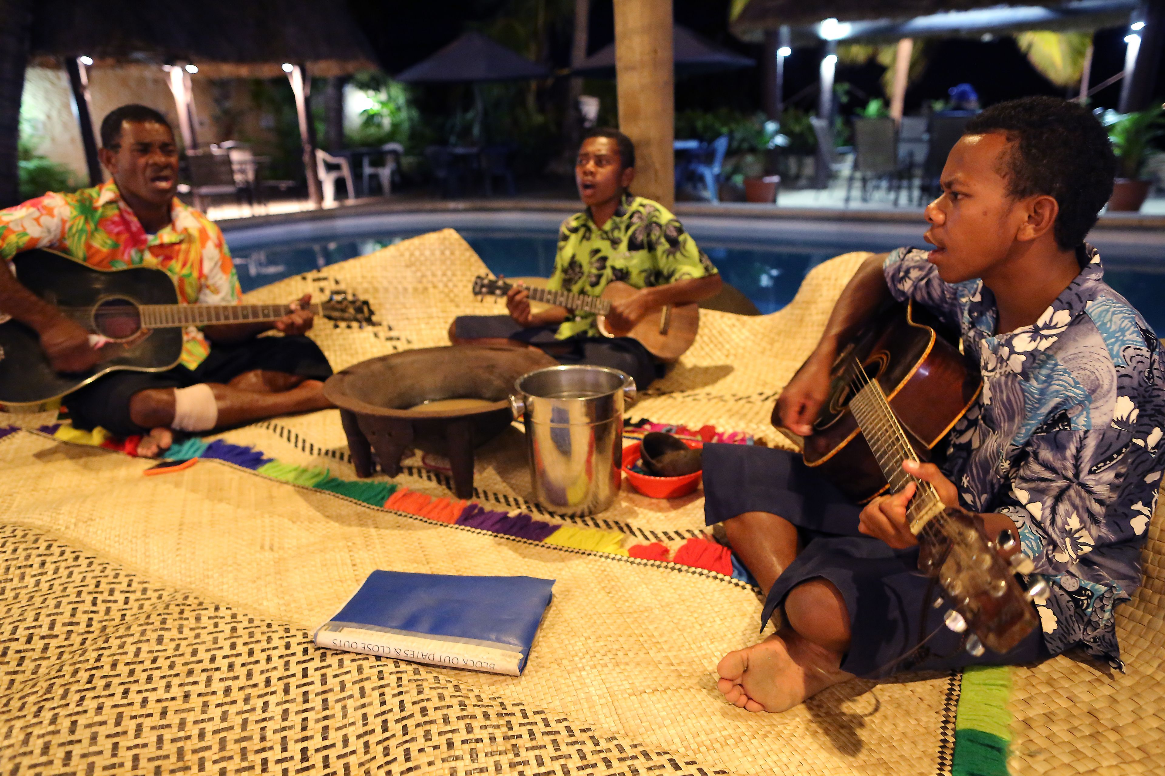 A group of people sitting on a woven mat by a poolside, playing guitars.