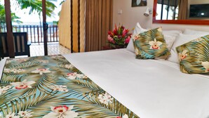 Une chambre d’hôtel avec un lit, des coussins à motifs floraux, une table de chevet ornée d’un vase de fleurs, et une vue sur les palmiers et l’océan.