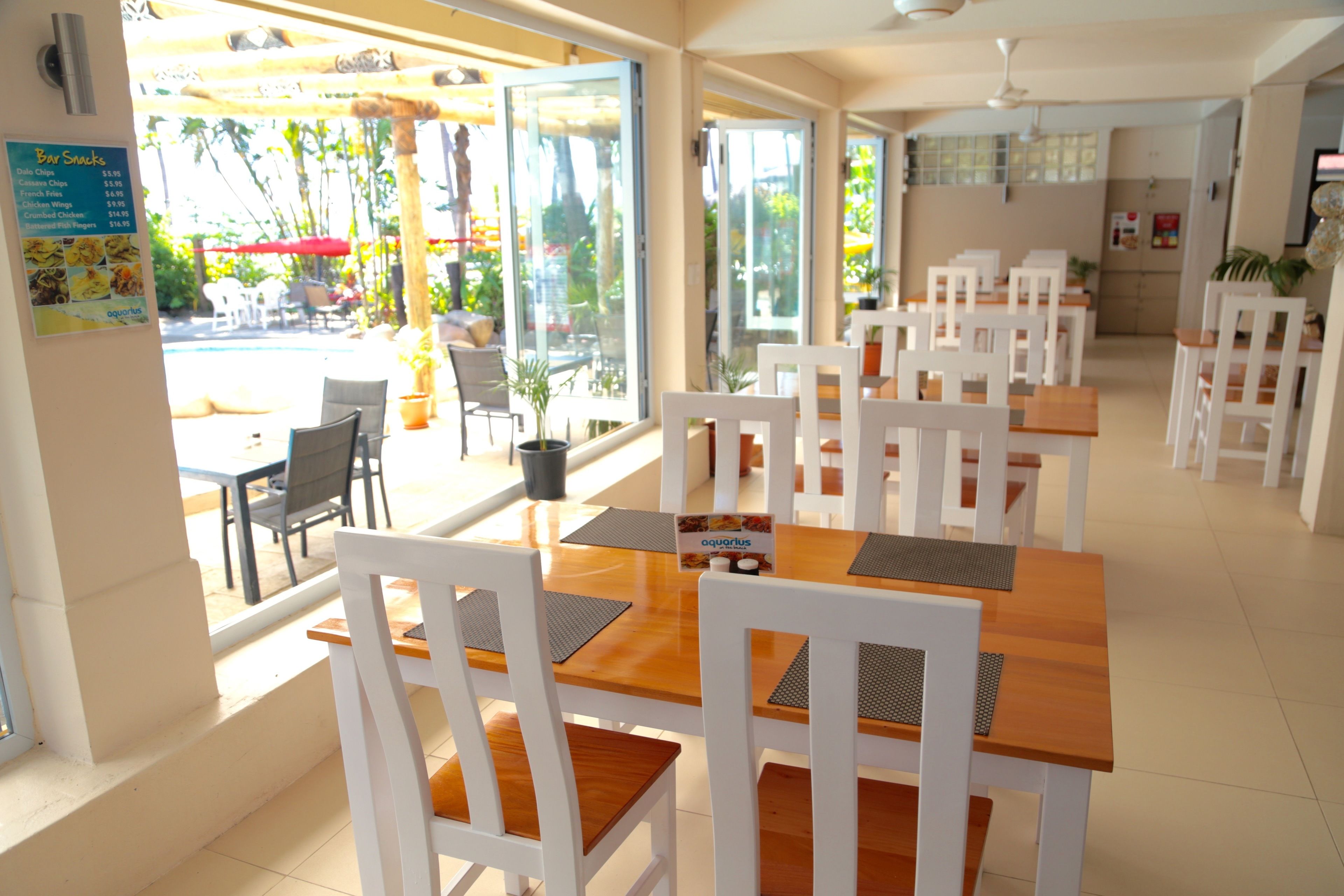 A restaurant with white chairs and wooden tables, large windows, and a menu board on the wall.