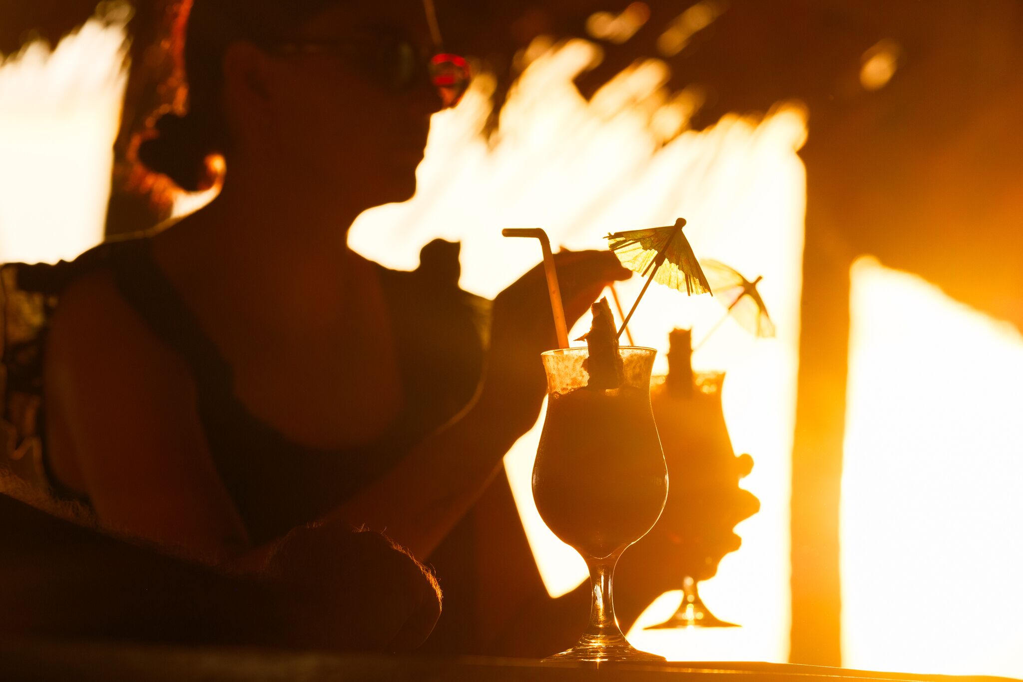 Une personne tenant un verre à cocktail avec une paille et un petit parasol, assise à une table, avec une lumière douce et chaleureuse en arrière-plan.