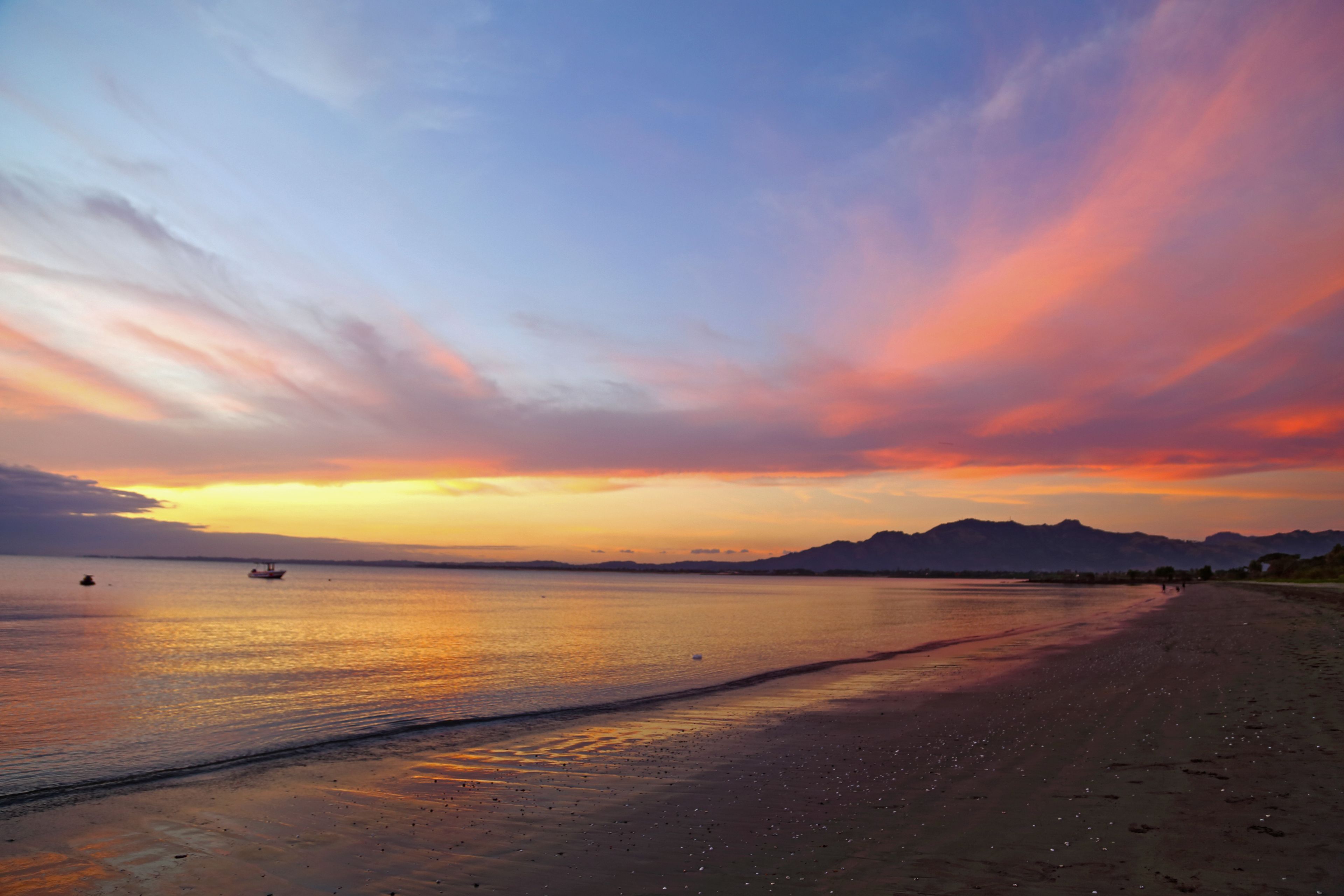 A beach with a calm sea, distant mountains, and a sunset sky.