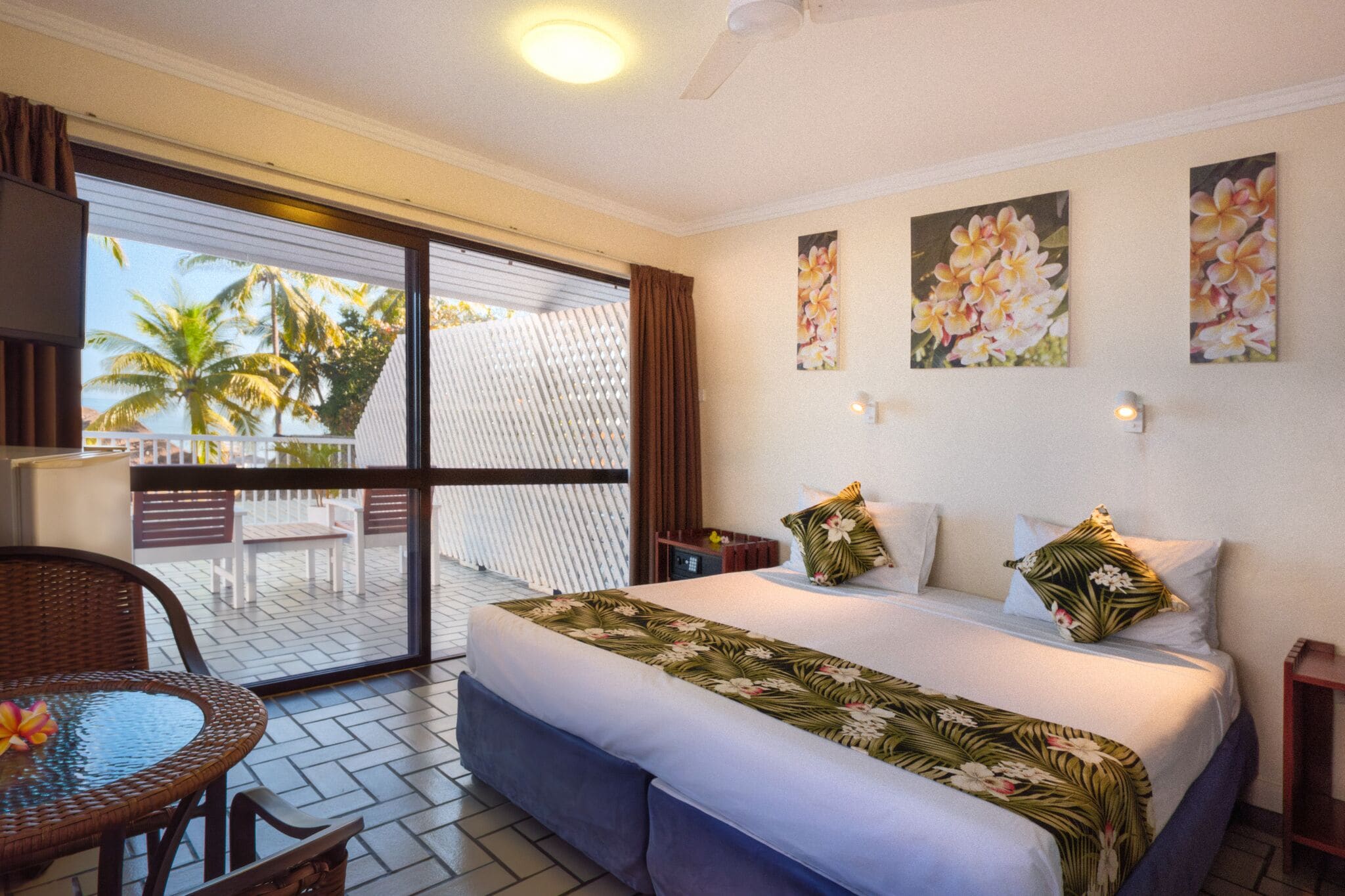 Habitación de hotel con una cama, una mesa y vistas a palmeras a través de una puerta corredera.