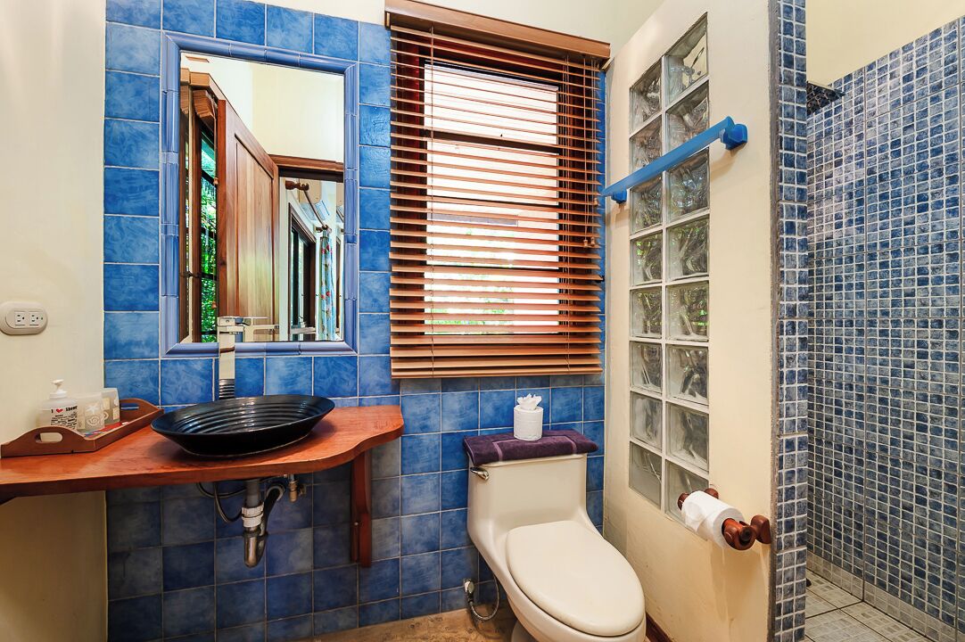 A bathroom with blue tiles, a wooden vanity, a toilet, and a window with blinds.