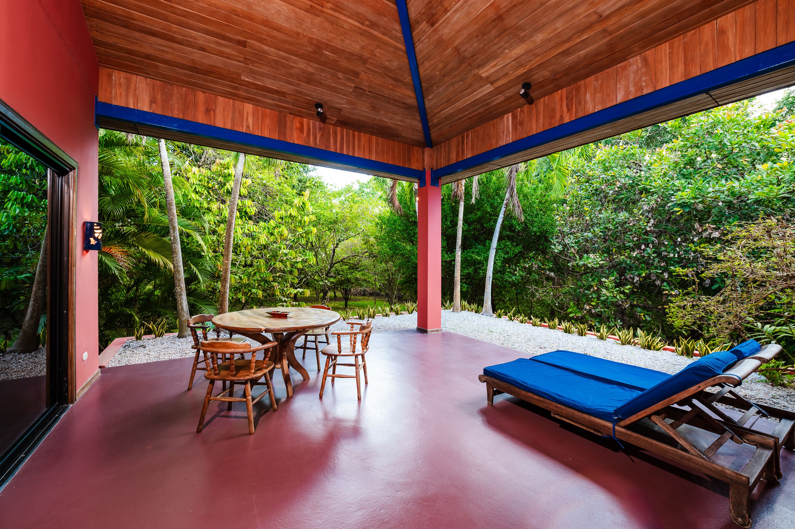 Ein überdachter Außenbereich mit Holztisch und -stühlen, einem blauen Lounge-Sessel und Blick auf üppiges Grün.