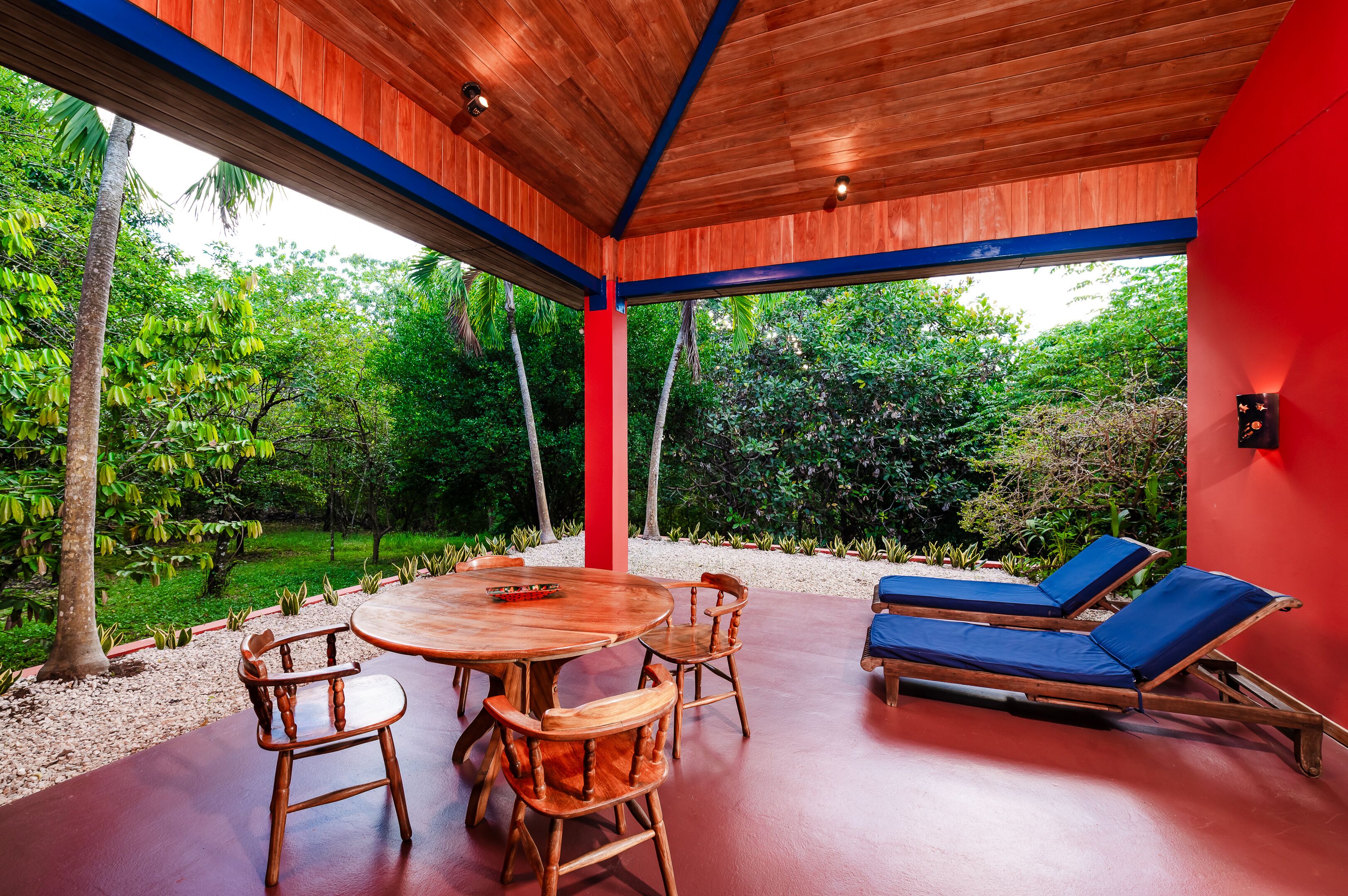 A covered outdoor area with a wooden table and chairs, a red wall, and a view of lush greenery.