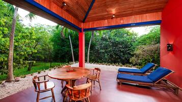 A covered outdoor area with a wooden table and chairs, a red wall, and a view of lush greenery.