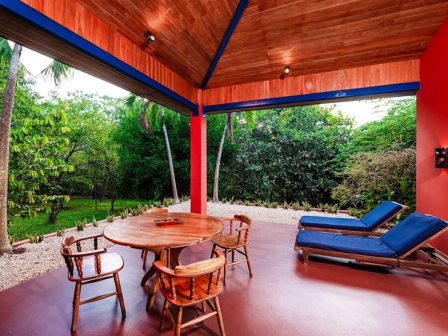 A covered outdoor area with a wooden table and chairs, a red wall, and a view of lush greenery.