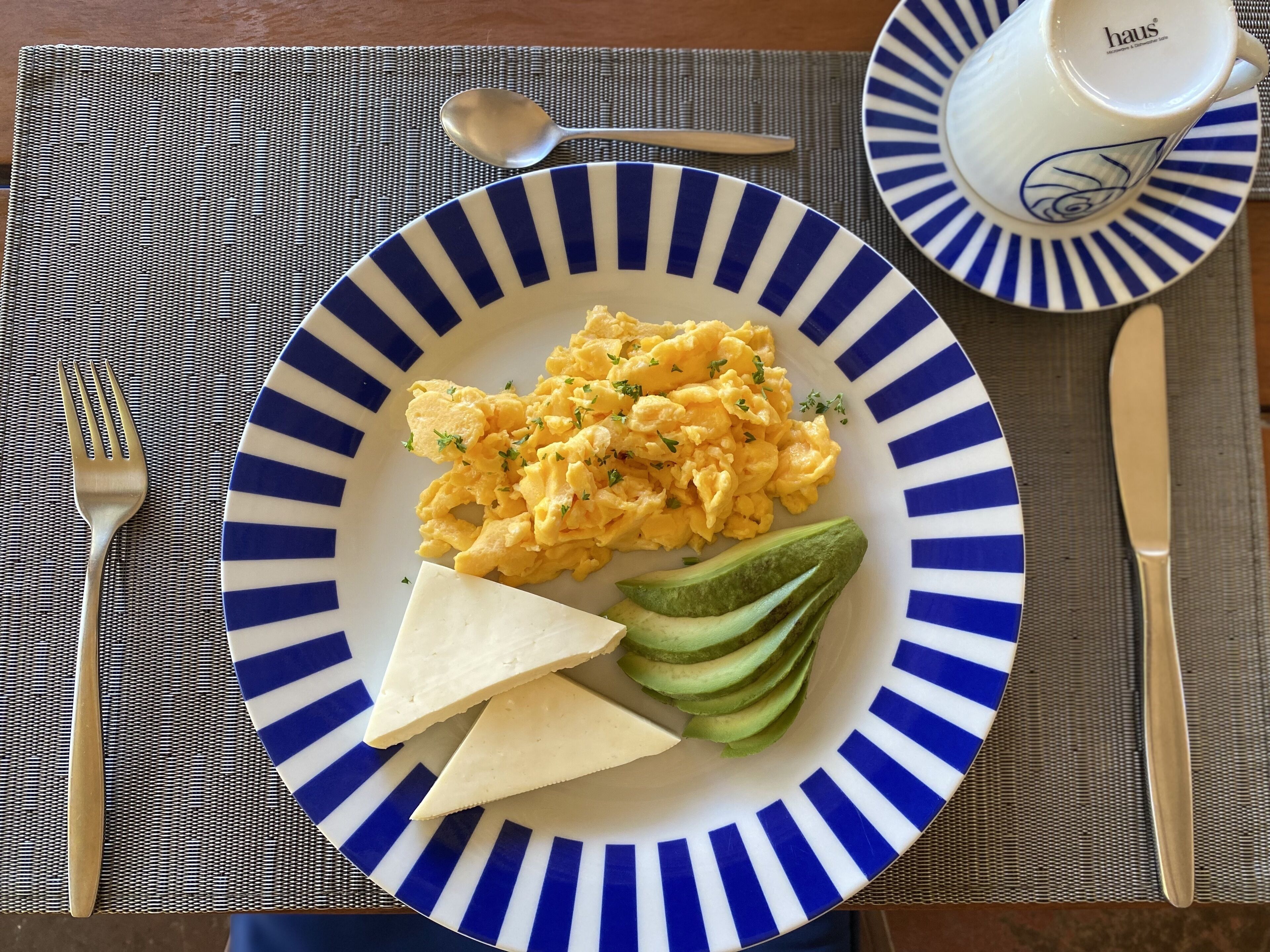 A plate with scrambled eggs, avocado slices, and cheese on a blue and white striped plate.