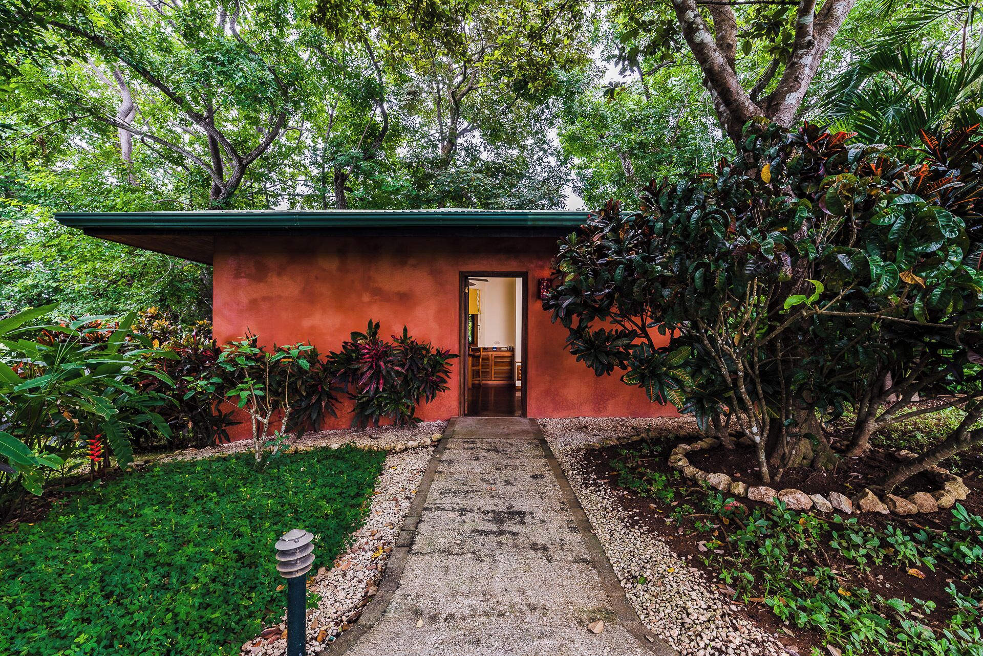 A small building with a red exterior and a green roof, surrounded by lush greenery and a gravel path leading to the entrance.