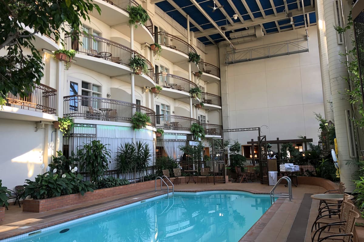 An indoor swimming pool with a clear view of the surrounding area, including balconies with railings and potted plants, and a seating area with tables and chairs.